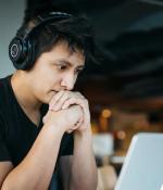 Photo of a man wearing over-ear headphones sitting at a table with his hands clasped together. He's staring at an open laptop in front of him.