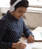Young woman sitting at laptop, wearing headphones, taking notes