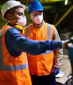 technician wearing reflective vest pointing at something while discussing results of joint work with his colleague