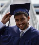 Young bearded man in graduation cap and gown smiles at the camera.